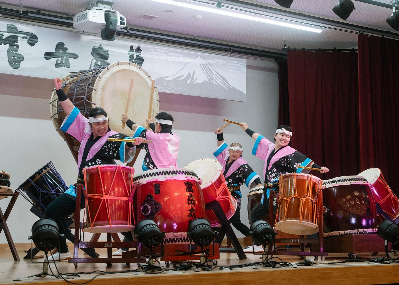 Taiko drummers in pink and black happi coats performing on stage, with a Mount Fuji banner behind them